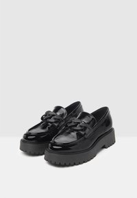 Mocassins en cuir verni noir avec une semelle compensée épaisse, ornés d'une chaînette décorative sur le dessus et d'une semelle texturée pour une meilleure adhérence.