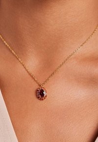 Gouden ketting met een ovale rode edelsteen, omringd door kleinere gekleurde stenen, die de ingewikkelde details en een glanzende afwerking benadrukt.