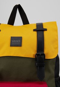 Sac à dos avec rabat jaune, sangle noire à boucle, corps vert olive, poche avant rouge et écusson logo Vans sur le rabat jaune.