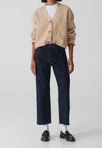 Cardigan beige a costine con bottoni, abbinato a jeans cropped blu scuro. Una T-shirt bianca sotto e mocassini marroni completano l'outfit.