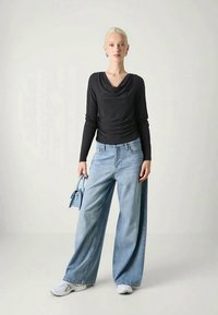 Black cowl-neck long sleeve top paired with wide-leg light blue jeans. Model holds a small blue handbag. White sneakers complete the outfit.
