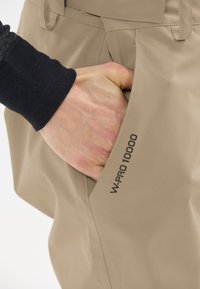 Pantalons étanches beiges avec une poche latérale zippée. Marque noire "W-PRO 10000" visible au niveau de la poche. Texture lisse.