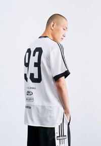 Balta sportinė marškinė su juodu numeriu "93" ir trimis juodomis juostelėmis ant rankovių. Pagaminta iš tinklinės tekstūros, su Adidas prekės ženklu ant apatinio krašto.