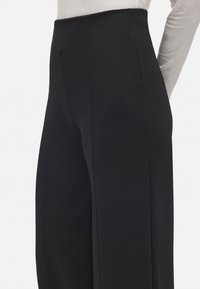 Primer plano de pantalones anchos de cintura alta en color negro combinados con una blusa de manga larga gris claro metida por dentro, sobre un fondo blanco.