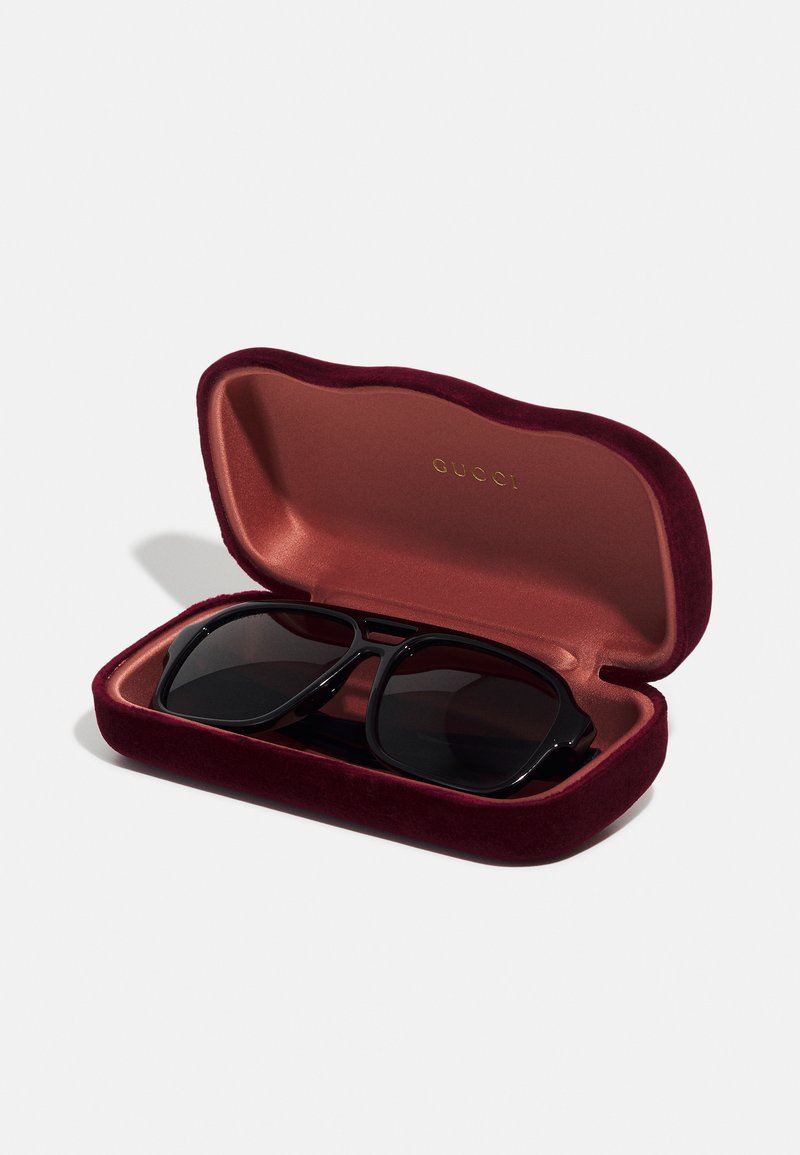 Lunettes de soleil noires avec une large monture à l'intérieur d'une boîte en velours bordeaux. La boîte a une texture douce et présente un logo doré à l'intérieur.