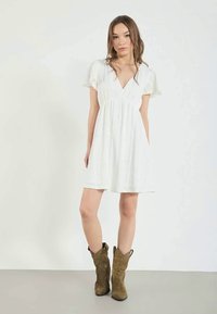 Vestido blanco corto con escote en V, mangas abullonadas y cintura fruncida. La modelo lleva botas vaqueras de color verde olivo. Textura suave, tejido ligero.