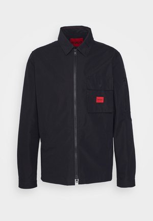 Schwarze Jacke mit einem Frontreißverschluss, einer Brusttasche mit Druckknopf und einem roten Etikett sowie einem Umlegekragen. Glatte Textur und lange Ärmel.