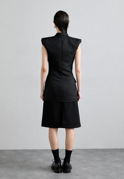 Priliehavé čierne šaty bez ruk�ávov so širokými ramennými panelmi, po kolená, viditeľné švy, skombinované s čiernymi ponožkami a robustnými topánkami.