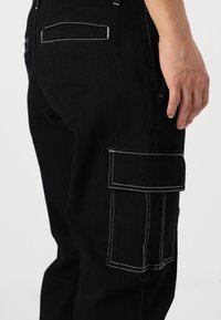 Črne denim cargo hlače z bočno našitno žepe, belim kontrastnim šivom in sproščenim krojem. Gladka tekstura, sproščen dizajn.