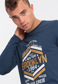 Marineblaues Langarm-T-Shirt mit einem großen grafischen Druck in Orange, Weiß und Blau, mit "Brooklyn" und zusätzlichem Text in fetter Schrift.