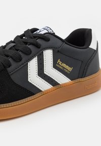 Baskets Hummel Handball Perfekt en noir et blanc avec semelle en caoutchouc, lacets noirs et bandes en chevron sur le côté en vue rapprochée.