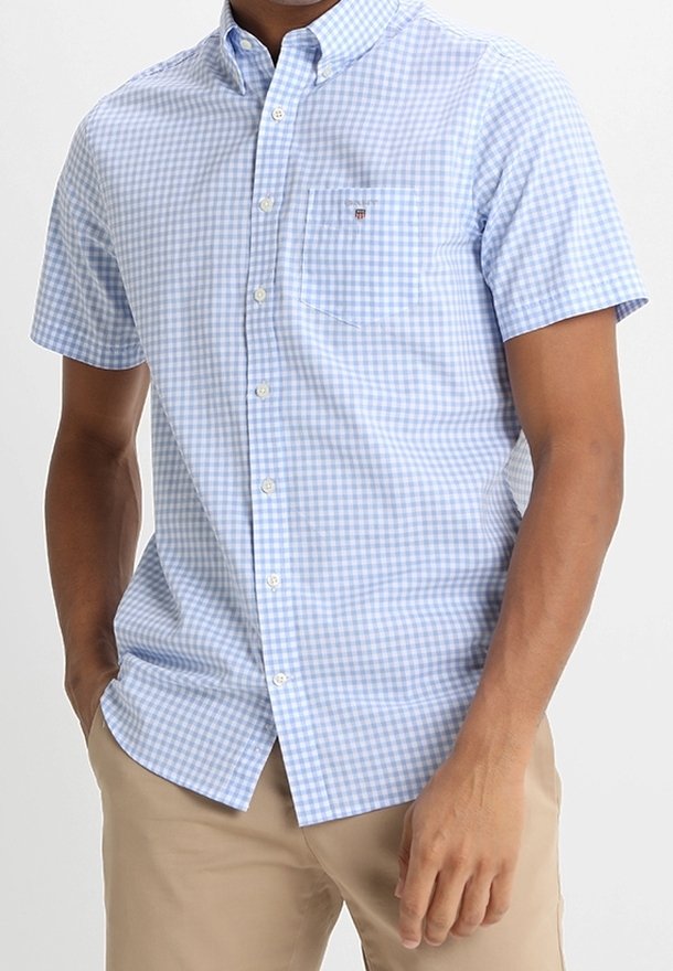 Homme portant une chemise à manches courtes à carreaux bleu clair et blanc avec un pantalon beige, debout avec une main dans la poche.