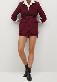 Burgundowa, strukturalna marynarka i krótka spódniczka w zestawie z białą koszulą z kołnierzykiem pod spodem; zawiera pasek do samodzielnego zawiązania i dopasowany krój. Czarne botki za kostkę.