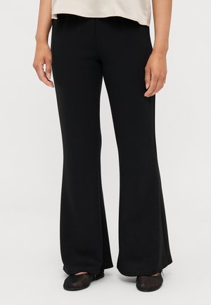 Vrouw draagt zwarte wijde broekspijpen en zwarte ballerina's, staand tegen een effen lichte achtergrond.