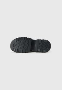 Suela de zapato negra con un profundo patrón de zigzag y formas geométricas sobre un fondo blanco liso.