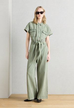 Kvinne iført lys grønn kortermet jumpsuit med knapper og knytt belte i midjen, svarte flate sko og svarte solbriller, står på tregulv.