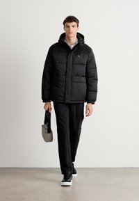 Homem a caminhar com uma jaqueta puffer preta, calças pretas, sapatilhas preto e branco, segurando uma pequena bolsa retangular cinza com uma alça preta.