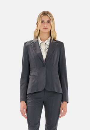 Blazer grigio aderente con chiusura a un bottone, due tasche frontali e cuciture dettagliate. Abbinato a una blusa bianca a fantasia.