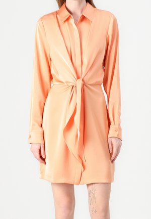 Femme portant une robe en satin pêche à manches longues avec un nœud sur le devant, debout devant un fond blanc uni.