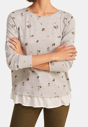 Frau mit verschränkten Armen, die ein hellgraues, mit Blumenmuster versehenes Langarmoberteil trägt, das über einem weißen Hemd und olivgrünen Hosen getragen wird.