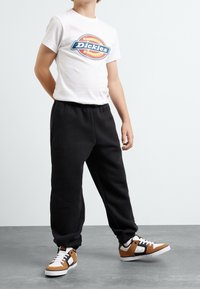 Vit t-shirt med blå och orange "Dickies" logga, ihop med svarta sweatpants och bruna, svarta och vita sneakers. Avslappnad ungdomsoutfit.