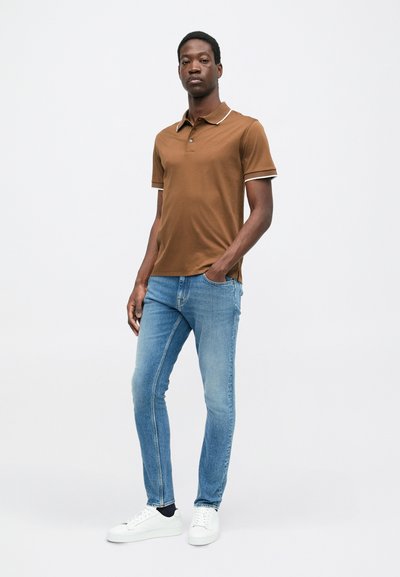 Hnědé polo tričko s bílým lemem, kombinované s světle modrými slim-fit džíny a bílými teniskami. Model stojí s rukama v kapsách.