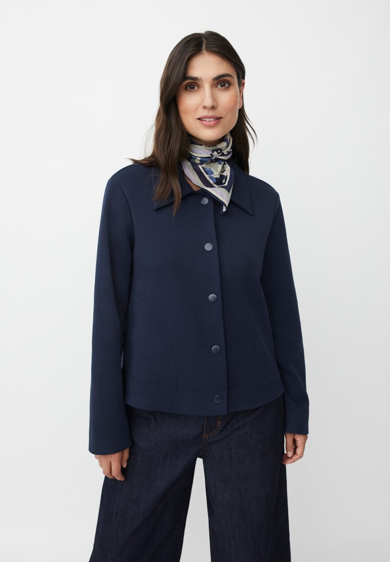 Kobieta o ciemnych włosach ubrana w granatową zapinaną na guziki kurtkę, ciemne szerokie jeansy i wzorzysty szalik związany na szyi, stojąca na tle białym.