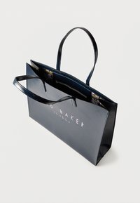 Τσάντα ώμου από γυαλιστερό δέρμα σε ναυτικό μπλε με μακριές λαβές, μεγάλη είσοδο και εσωτερικό με floral σχέδιο. Διαθέτει ανάγλυφο λογότυπο στο μπροστινό μέρος.