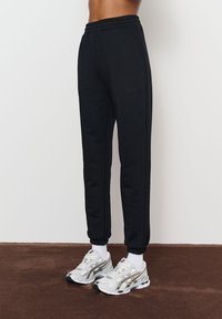 Pantaloni sport negri cu tivuri elasticizate, prevăzuți cu buzunare laterale și un logo discret al brandului pe față. Asortați cu adidași albi.