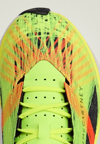 Chaussure de sport avec un empeigne jaune vif ornée de fils multicolores, des accents noirs et des détails perforés. Surface texturée et lacets inclus.