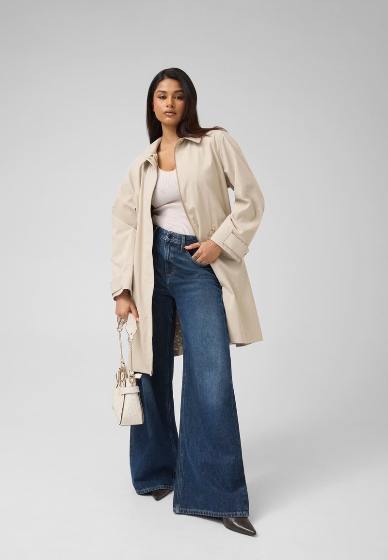 Femme portant un trench beige, un top blanc, un jean bleu à jambes larges et des chaussures noires pointues, tenant un sac à main blanc sur un fond uni.