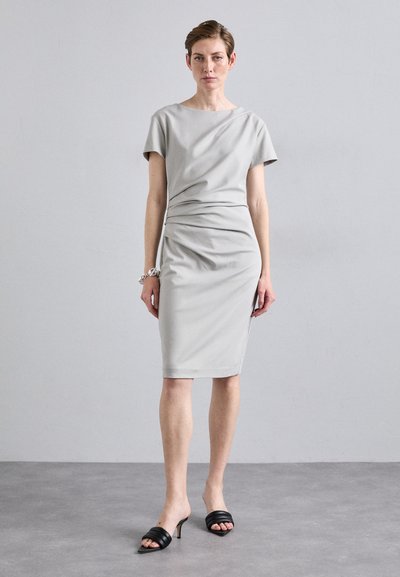 Robe grise ajustée avec des manches courtes, froncée à la taille. Tissu lisse, longueur genou. Associée à des mules noires à talons. Fond uni.