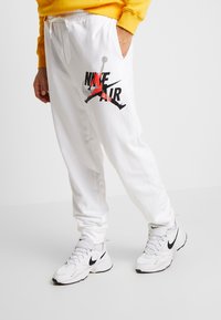 Bílé teplákové kalhoty mají výrazný černý a červený nápis „NIKE AIR“. Jsou vyrobeny z měkkého materiálu, mají elastický pas a boční kapsy.