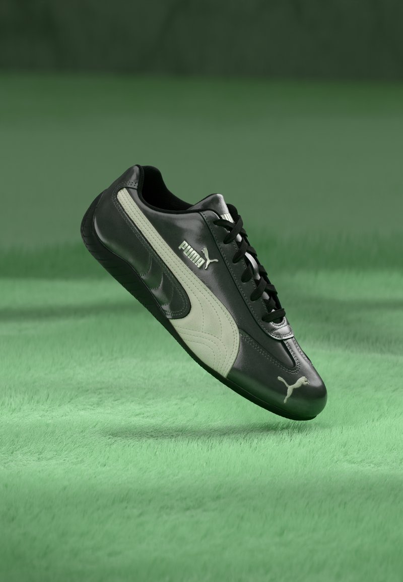 Musta-valkoinen Puma-tossu riippuu vihreän tekstuuripinnan yllä, näyttäen selvästi sivun, jossa on Puma-logo ja nauhat.