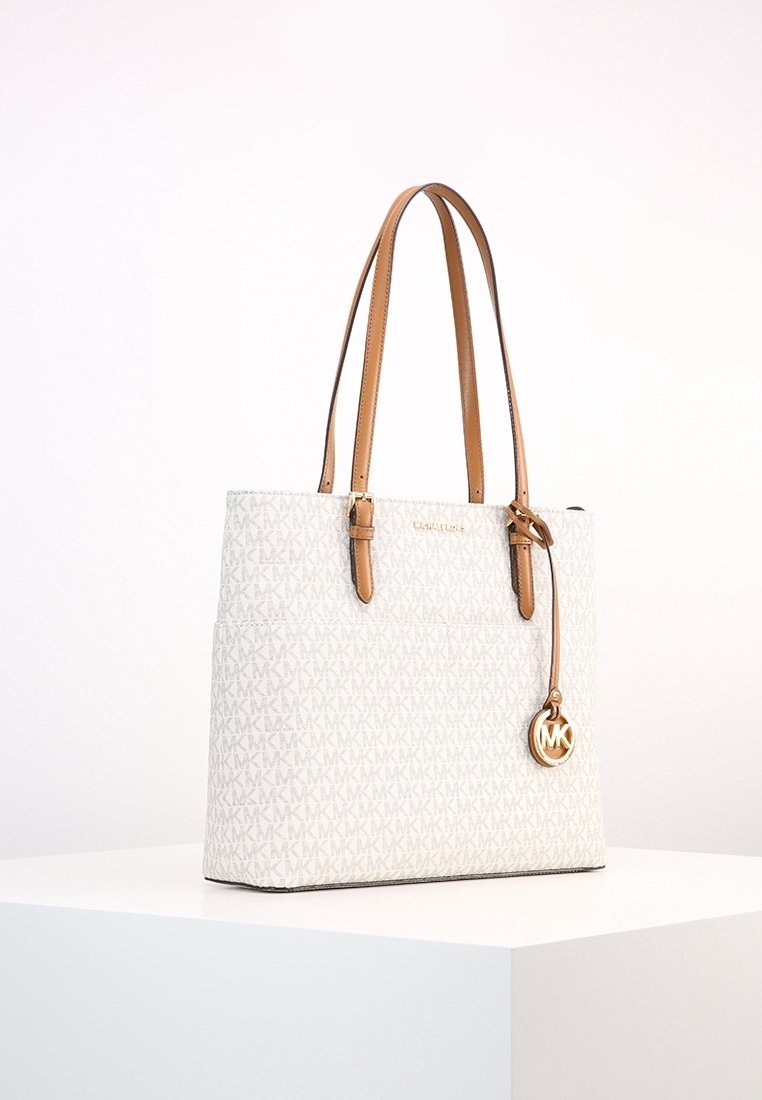 Sac cabas blanc Michael Kors avec poignées en cuir beige et breloque logo dorée exposé sur un socle blanc.