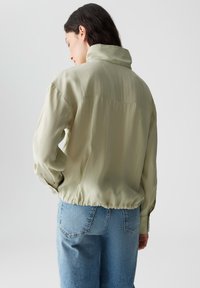 Lichtgroene, loose-fitting jas met een hoge kraag, gathered zoom en een zachte textuur. Gedragen over high-waisted, lichtblauwe denim jeans.