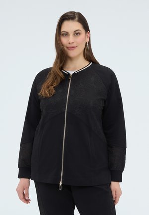 Femme portant une veste noire zippée avec des motifs floraux sur la poitrine et les manches, debout devant un fond clair uni.