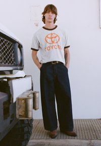 Sivá tričko s oranžovým logom „TOYOTA“, čiernym lemom; voľné tmavé džínsy; stojí vedľa vozidla s textúrovanou čiernou pneumatikou.