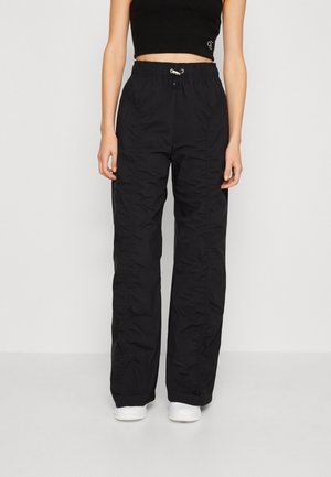 Persona con pantalones negros sueltos con cintura elástica y zapatillas blancas, mostrada desde el cuello hasta los pies sobre un fondo liso.
