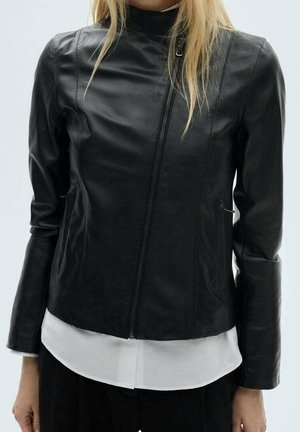 Mujer con una chaqueta de cuero negra ajustada con cierre diagonal sobre una camisa blanca, vista desde los hombros hasta las caderas contra un fondo liso.