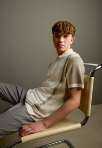 Īsām piedurknēm polo krekls krēmkrāsā ar gaiši brūnām vertikālām svītrām, piedurknēm ar ribveida tekstūru, kopā ar gaismas pelēkām biksēm, sēžot krēslā.