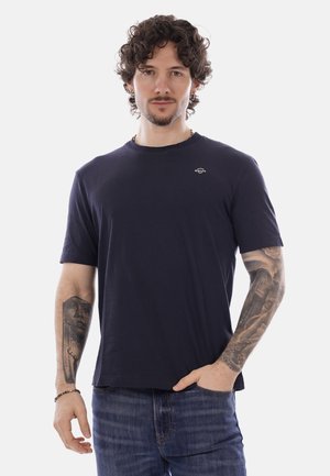 Uomo con capelli ricci e tatuaggi che indossa una maglietta blu navy e jeans, posando con una mano in tasca su sfondo bianco.