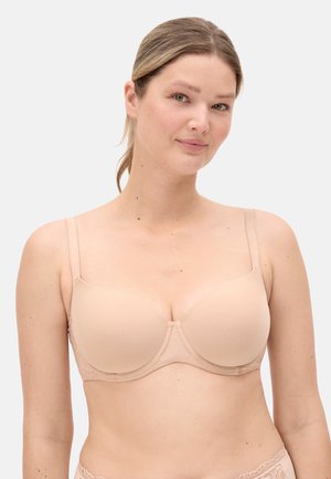 Mujer con un sujetador acolchado color nude con tirantes ajustables, de pie frente a un fondo liso gris claro, mirando hacia adelante con una leve sonrisa.