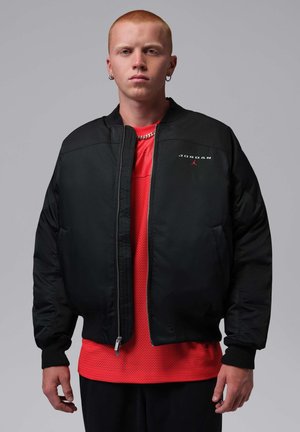 Junge Person mit kurzen roten Haaren trägt eine schwarze Jordan-Jacke über einem roten Sportshirt vor grauem Hintergrund.