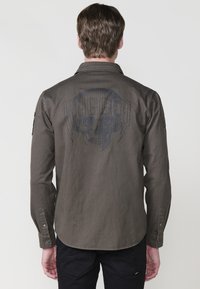 Olijfgroene long-sleeve shirt met schedelgrafiek en de tekst "RUGGED". Voorzien van knoopsluiting bij de mouwen en een gebogen onderkant. Van katoenmix materiaal.