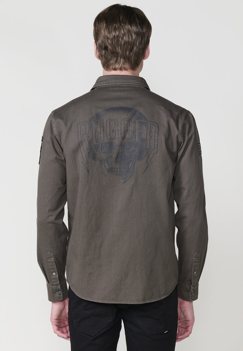Olijfgroene long-sleeve shirt met schedelgrafiek en de tekst "RUGGED". Voorzien van knoopsluiting bij de mouwen en een gebogen onderkant. Van katoenmix materiaal.