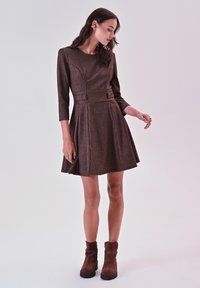 Bronzen ribbel jurk met driekwart mouwen, aangesloten taille, A-lijn rok en knoopaccenten. Combinatie met bruine enkellaarzen.