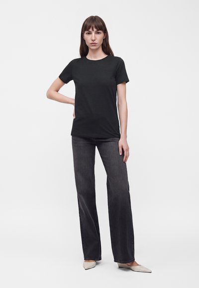 Femme debout, une main sur la hanche, portant un t-shirt noir uni, un jean foncé à jambes larges et des chaussures claires pointues sur fond blanc.