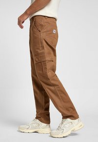 Pantalones cargo marrones con un corte recto, múltiples bolsillos laterales y detalles de costura. Combinados con zapatillas deportivas de color claro.