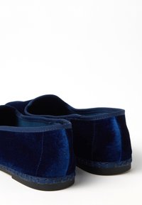 Un paio di scarpe slip-on in velluto blu scuro con suole nere, viste dal retro su una superficie bianca.